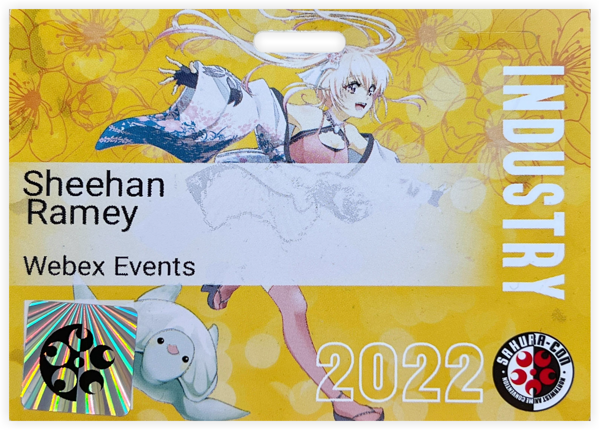 A pre-printed badge with an anime character running on a yellow background with floral designs. Attendee information is printed on a lighter section near the middle of the badge.
